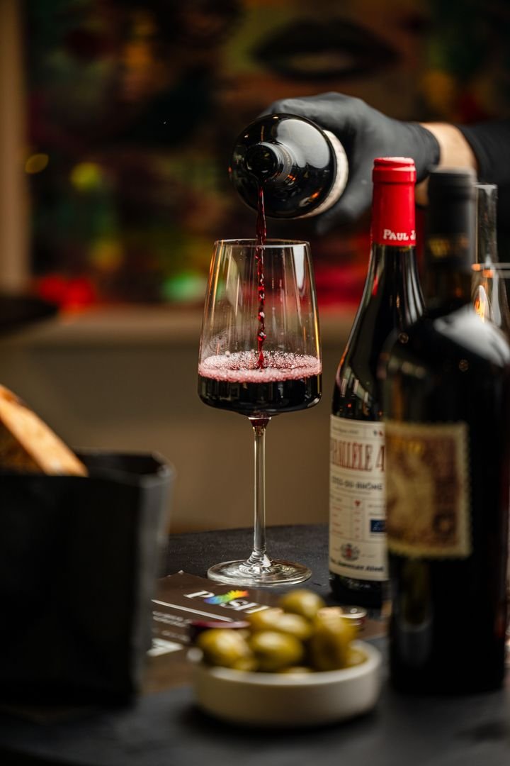 Rotwein wird mit schwarzem Handschuh in ein Glas eingeschenkt, daneben Oliven